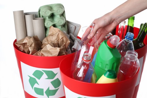 The image displays two red recycling bins with white recycling symbols on their sides, placed side by side on a plain white background. The left bin contains several vertically standing cardboard tubes, crumpled brown paper, and an egg carton made of light green molded pulp. The right bin is filled with transparent plastic bottles, some with coloured caps and labels, and a green plastic container with a red cap. A person's hand, wearing a clear plastic glove, is seen reaching into the right bin, possibly for sorting or removing items. The setting appears to be a controlled environment for waste sorting or recycling preparation. This scene illustrates general rubbish collection and recycling activities, relevant to waste management and clearance services in urban environments such as Covent Garden, with a focus on recyclable materials like paper, cardboard, and plastics.